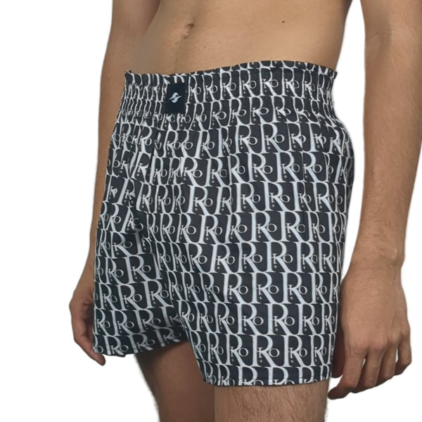 Person wearing black swim shorts with white letter pattern on a plain background